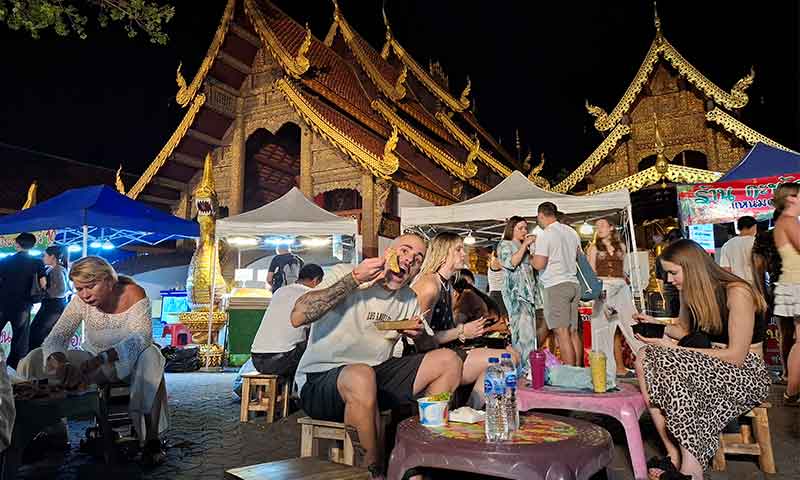 Turistas cenando en las mesas del mercado nocturno junto al templo Wat Phra Singh iluminado de noche en Chiang Mai